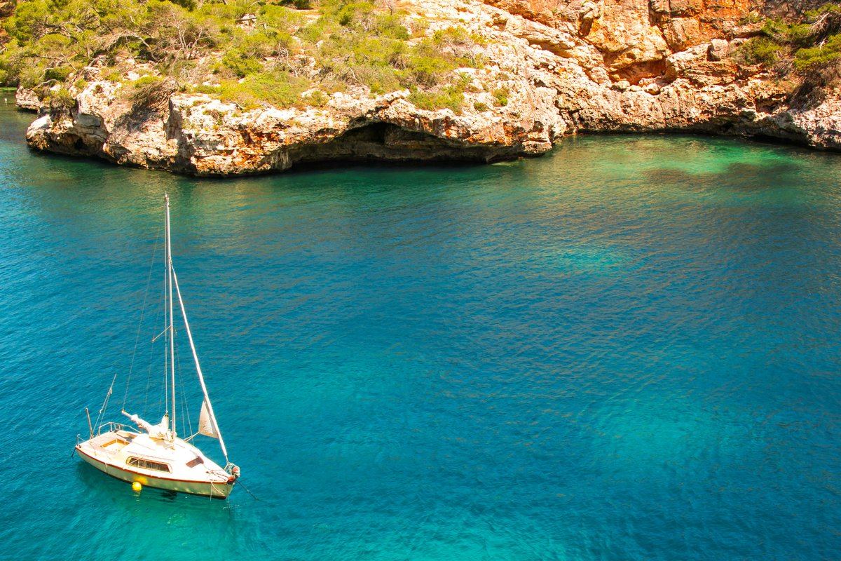 A sailboat at anchor in a turquoise cove below limestone cliffs, Mallorca